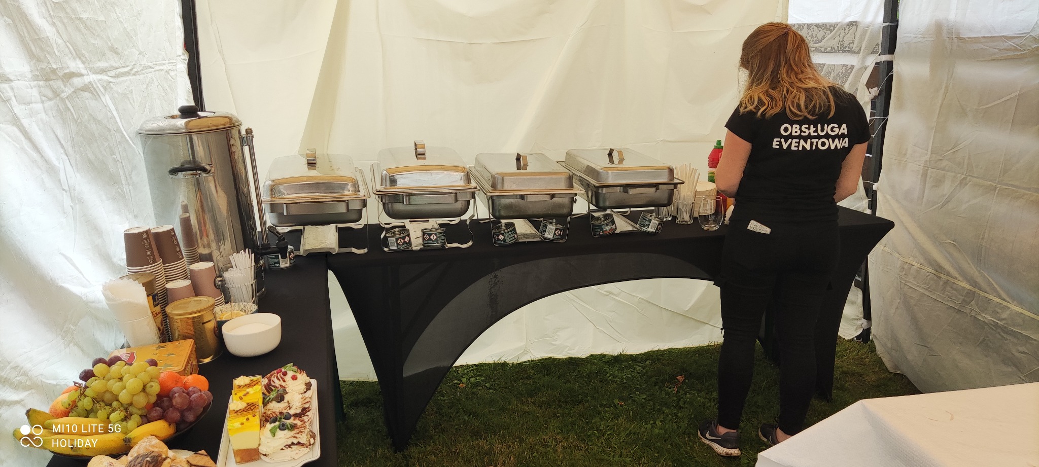 Stół z podgrzewaczami do potraw, owocami i deserami w namiocie, obsługa eventowa w tle.