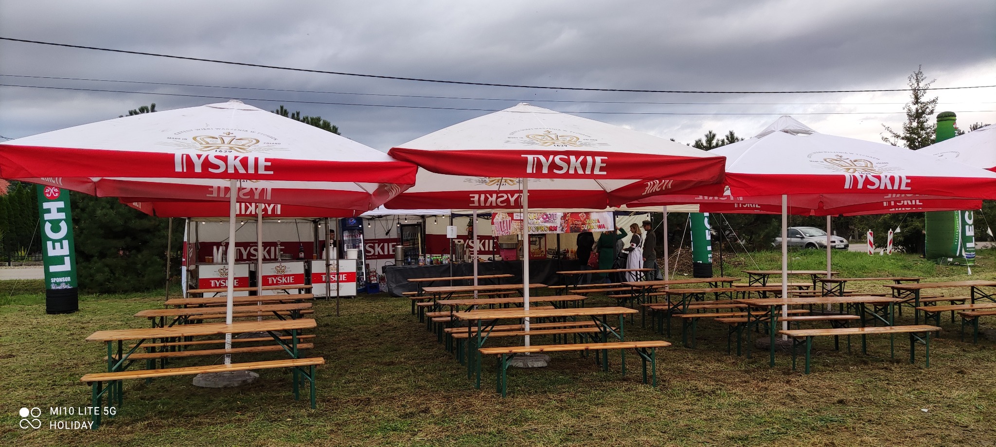Plenerowa strefa gastronomiczna z rzędami drewnianych stołów i ławek, osłonięta białymi parasolami z czerwonymi pasami i logo Tyskie, widoczne stoiska z napojami i przekąskami, w tle pochmurne niebo.