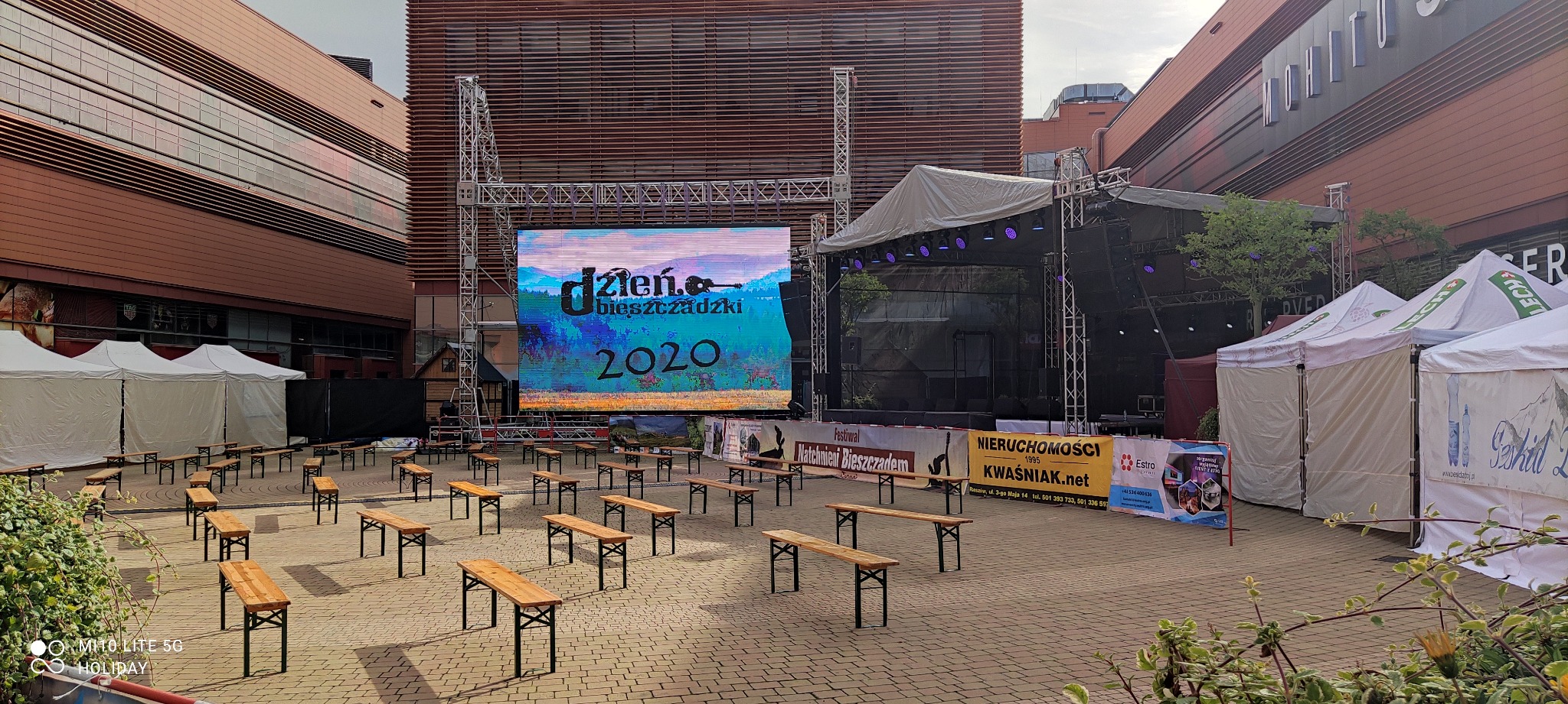 Przygotowania do plenerowej imprezy: scena z telebimem wyświetlającym nazwę wydarzenia 'Dzień Bieszczadzki 2020', rzędy drewnianych ławek dla publiczności, białe namioty w tle, budynki w tle.