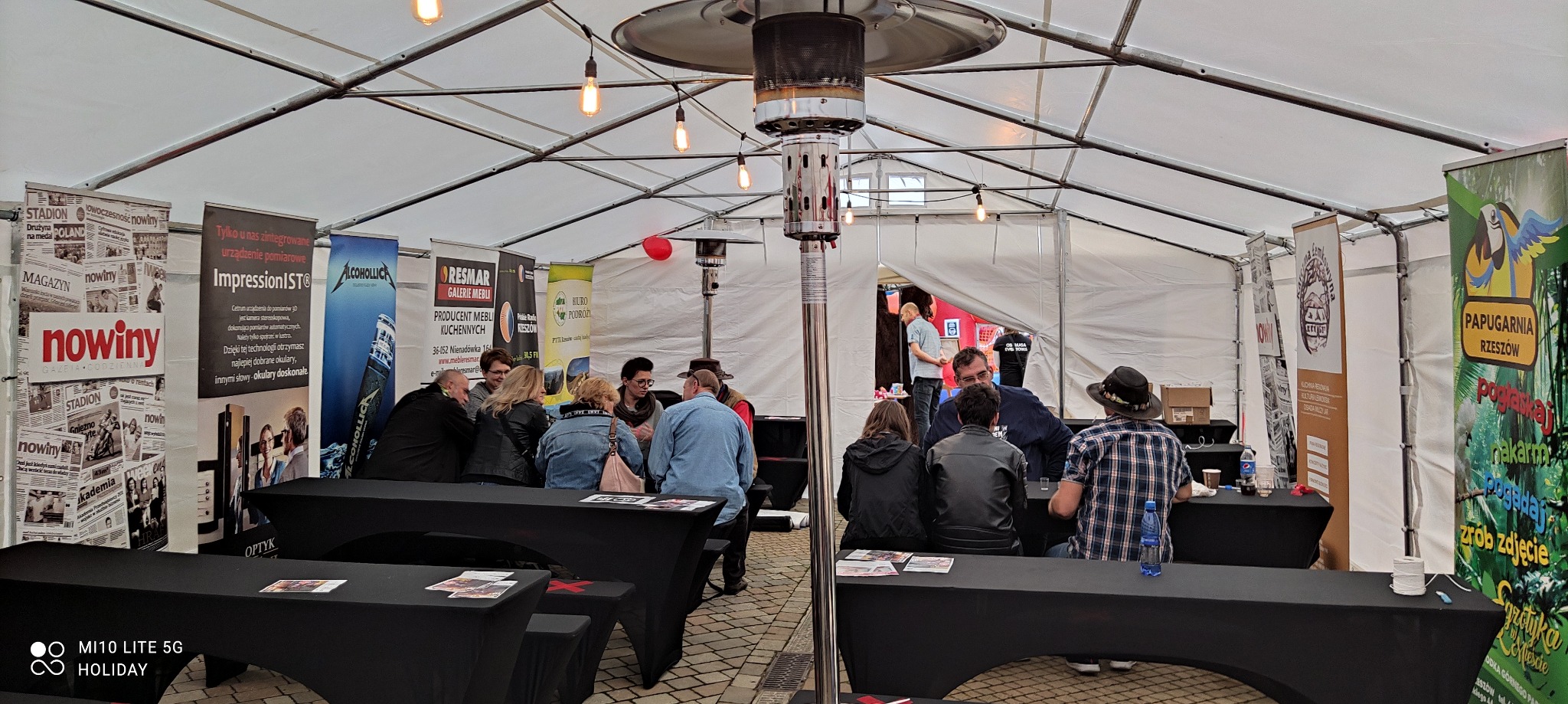 Wnętrze białego namiotu eventowego z metalowym ogrzewaczem, podwieszonymi żarówkami i stolikami, przy których siedzą goście, w tle widoczne roll-upy reklamowe różnych firm.