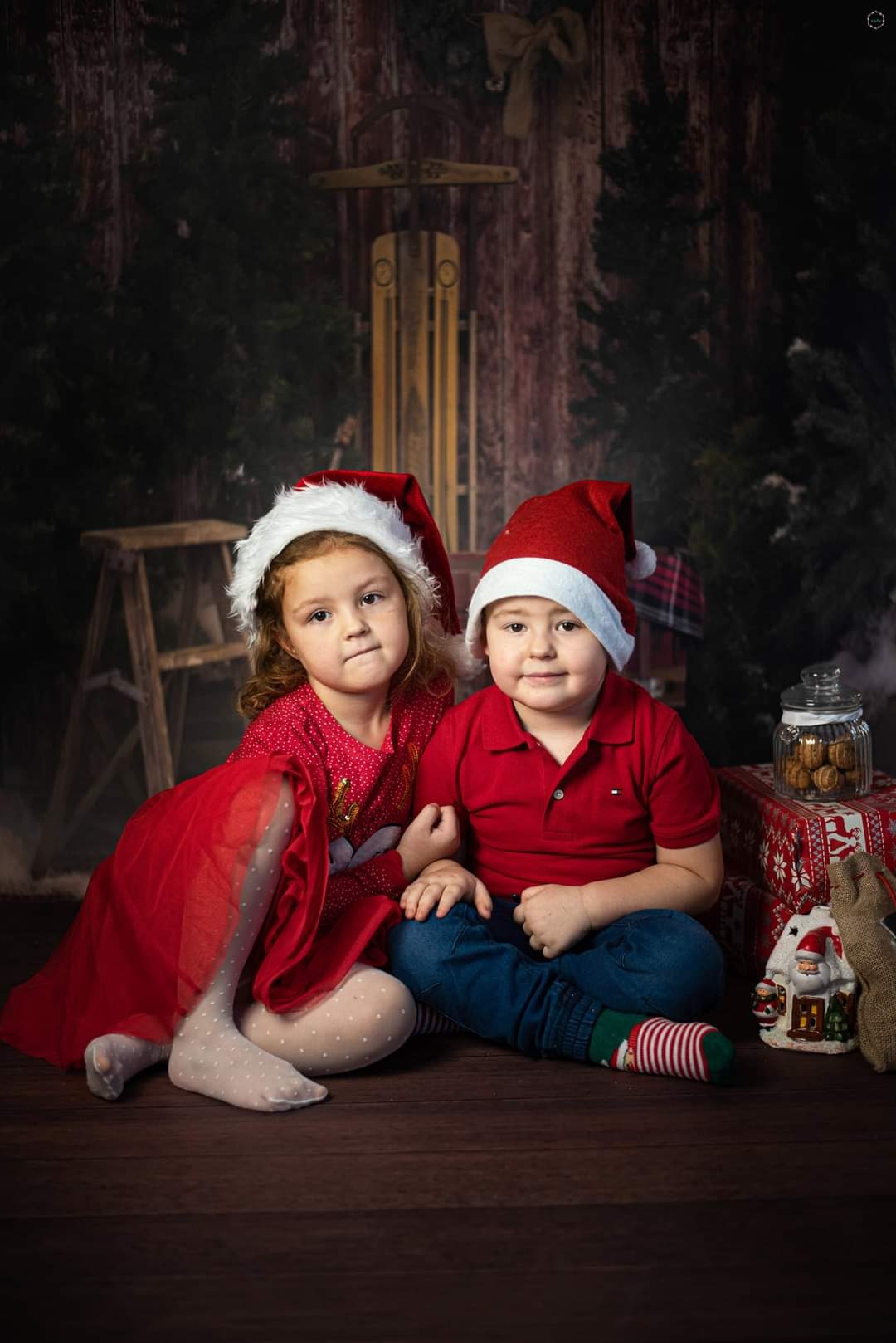 Dwoje dzieci w czapkach Mikołaja pozuje do zdjęcia w Grudziądzu, siedząc na podłodze przed świątecznym tłem z saniami i prezentami.
