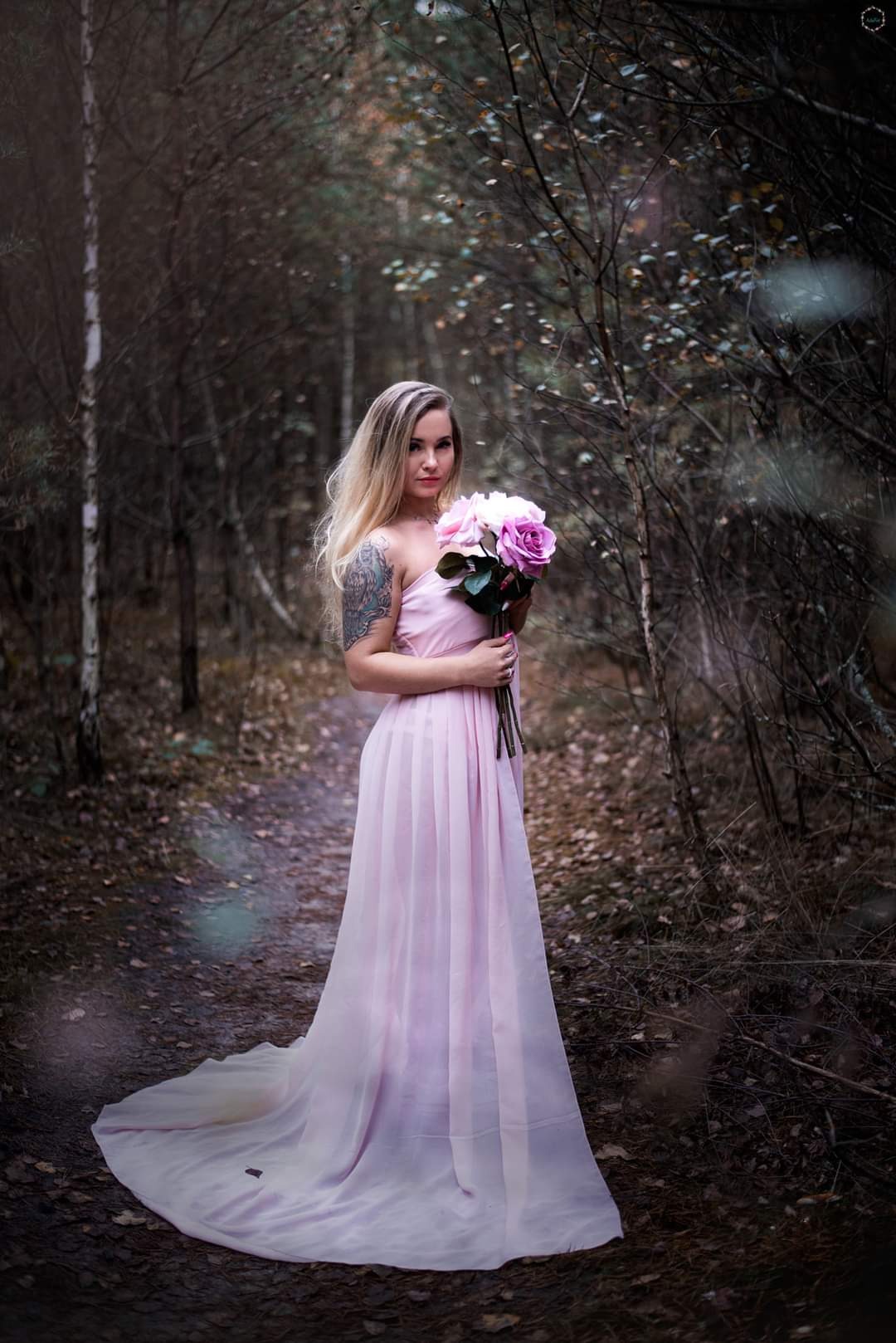 Kobieta w zwiewnej, pudrowo-różowej sukni trzyma bukiet róż w leśnej scenerii, z widocznym tatuażem na ramieniu i delikatnym makijażem.