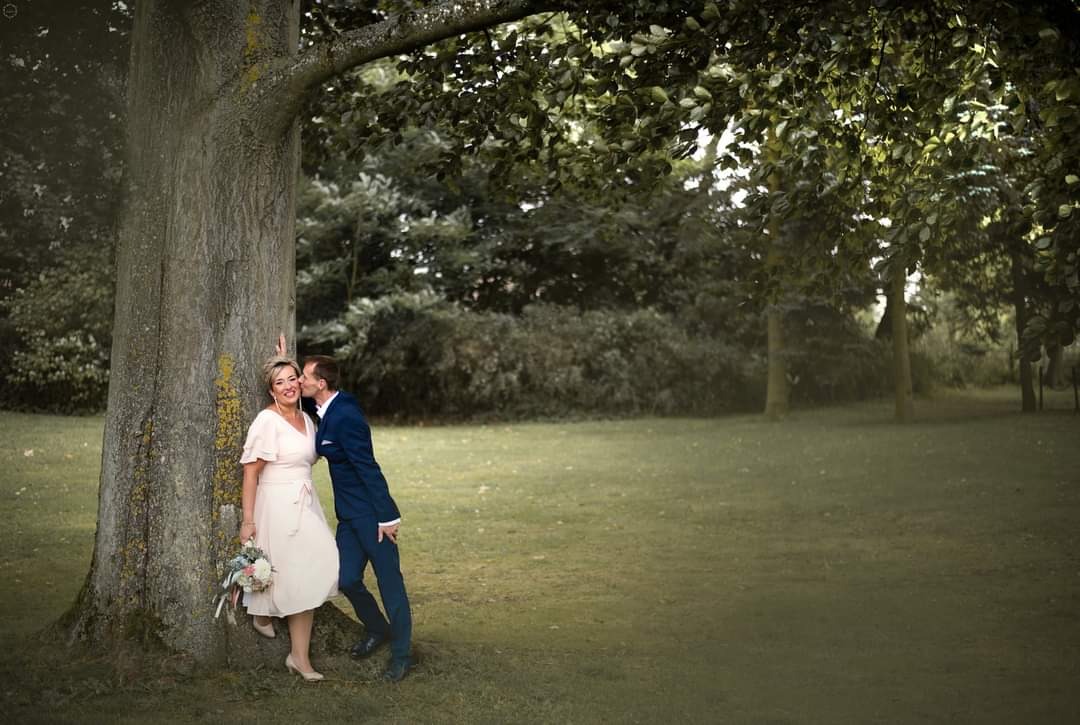 Uśmiechnięta kobieta w jasnej sukience z bukietem kwiatów opiera się o drzewo, mężczyzna w granatowym garniturze całuje ją w policzek na tle zielonego parku.