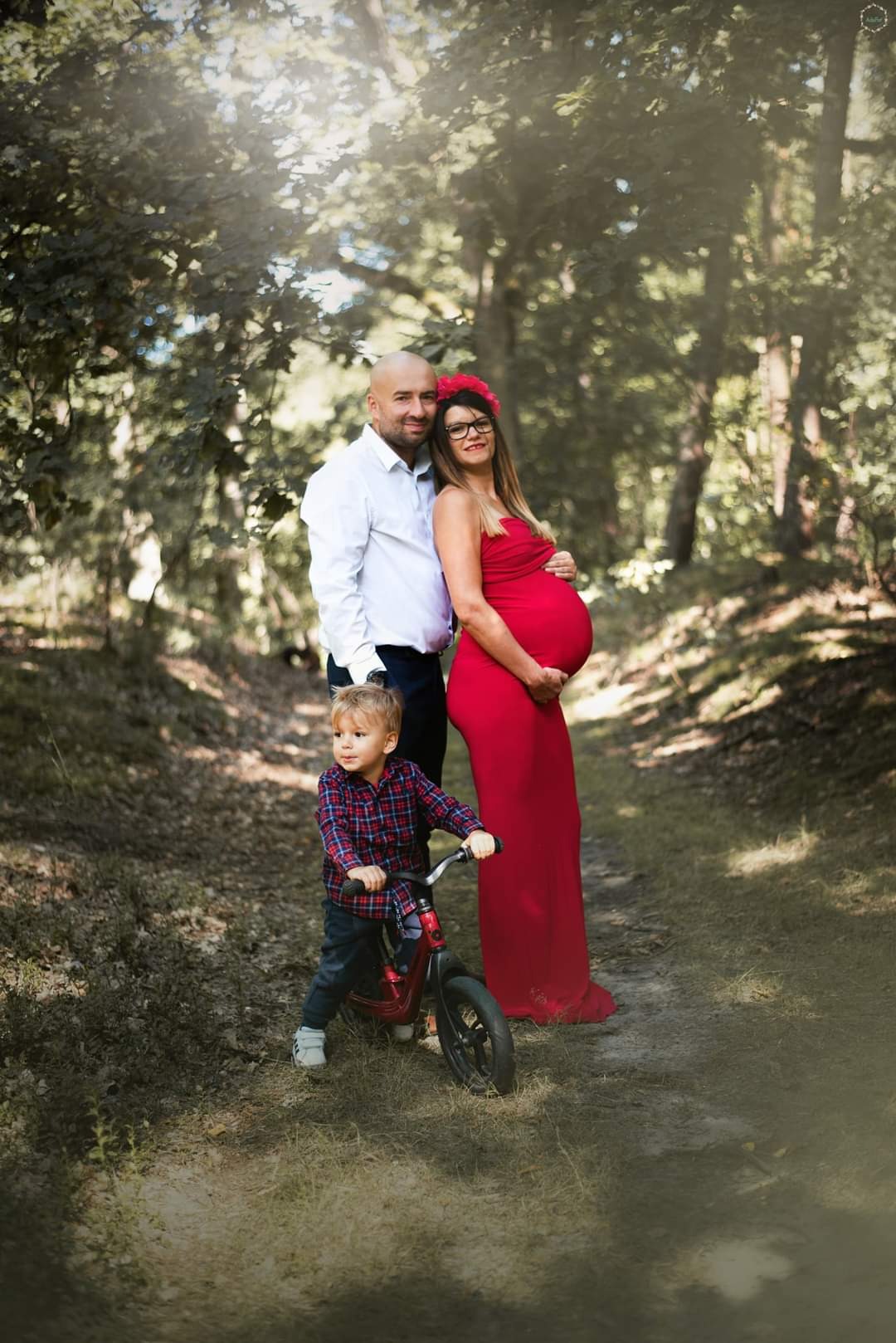 Ciężarna kobieta w czerwonej sukni, mąż i syn na rowerku w leśnej scenerii, delikatne światło słoneczne.