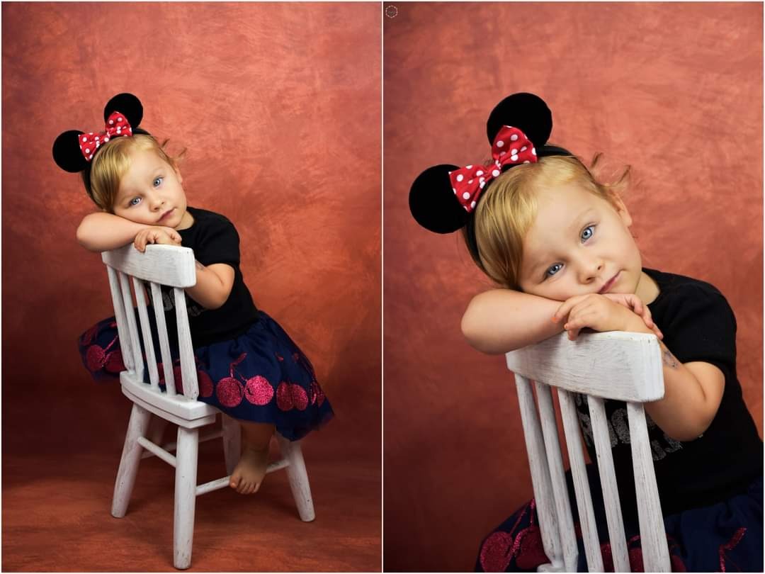 Dziewczynka z blond włosami i niebieskimi oczami, ubrana w czarną koszulkę i granatową spódniczkę w wiśnie, pozuje na białym krześle, opierając głowę o oparcie. Ma na głowie opaskę z uszami Myszki...