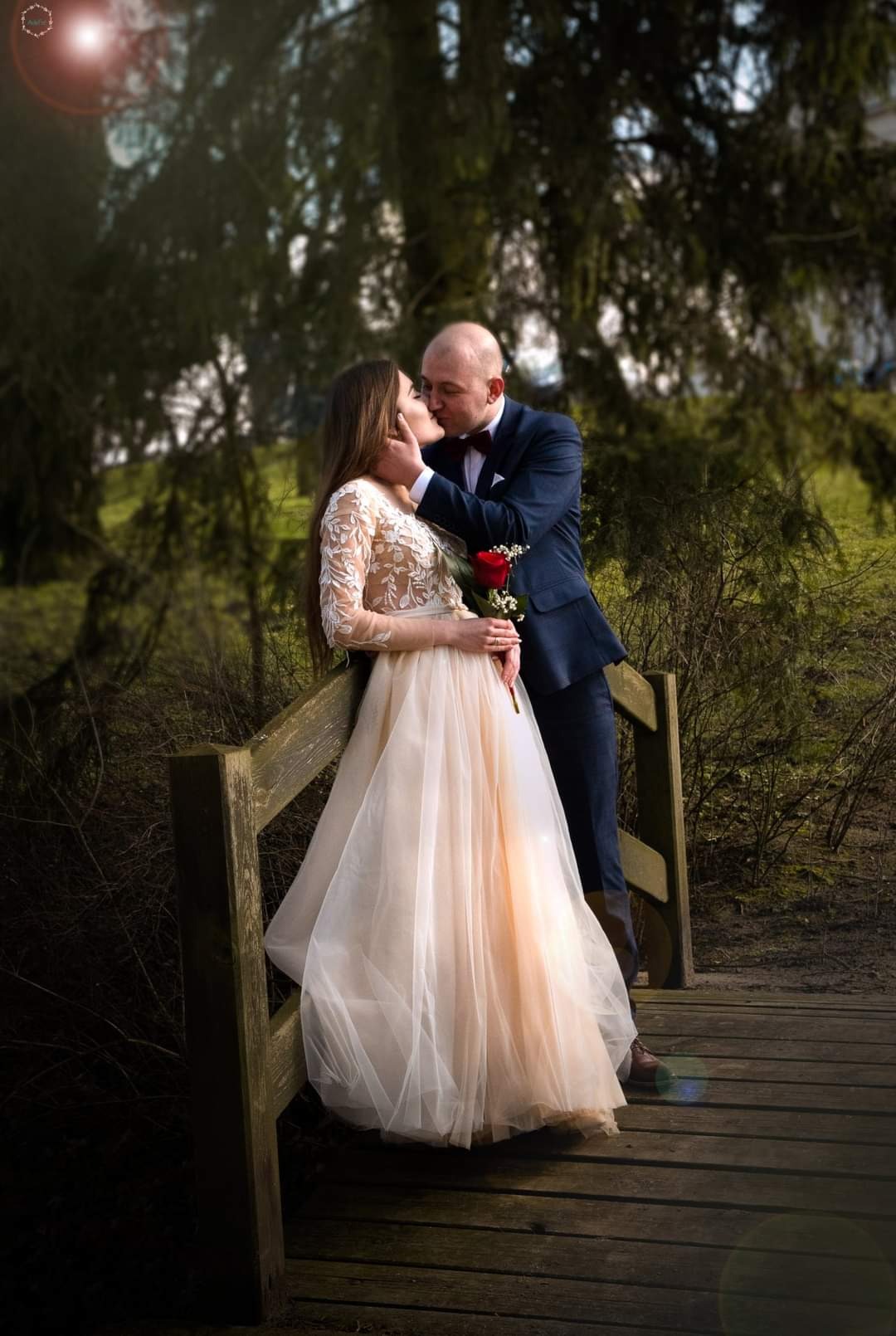 Para całująca się na drewnianym moście w plenerze, panna młoda w zwiewnej sukni z koronkowymi rękawami trzyma czerwoną różę, pan młody w granatowym garniturze z muchą.