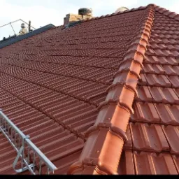 Czerwona dachówka ceramiczna na skośnym dachu z zamontowaną aluminiową barierką zabezpieczającą, widok na komin w tle.