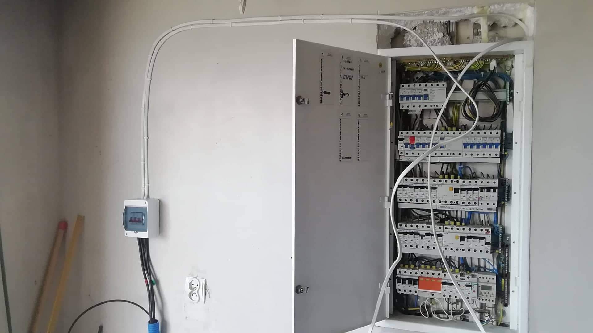 Otwarta skrzynka elektryczna z widocznymi bezpiecznikami i przewodami, poprowadzona instalacja w białych listwach na ścianie, surowy stan wykończenia wnętrza.