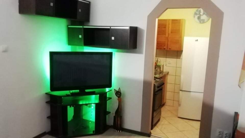 Telewizor z podświetleniem LED w kolorze zielonym, zamontowany na czarnej szafce RTV z półkami, obok drewniana figurka kota, na ścianie wiszące szafki, widok na kuchnię w tle.