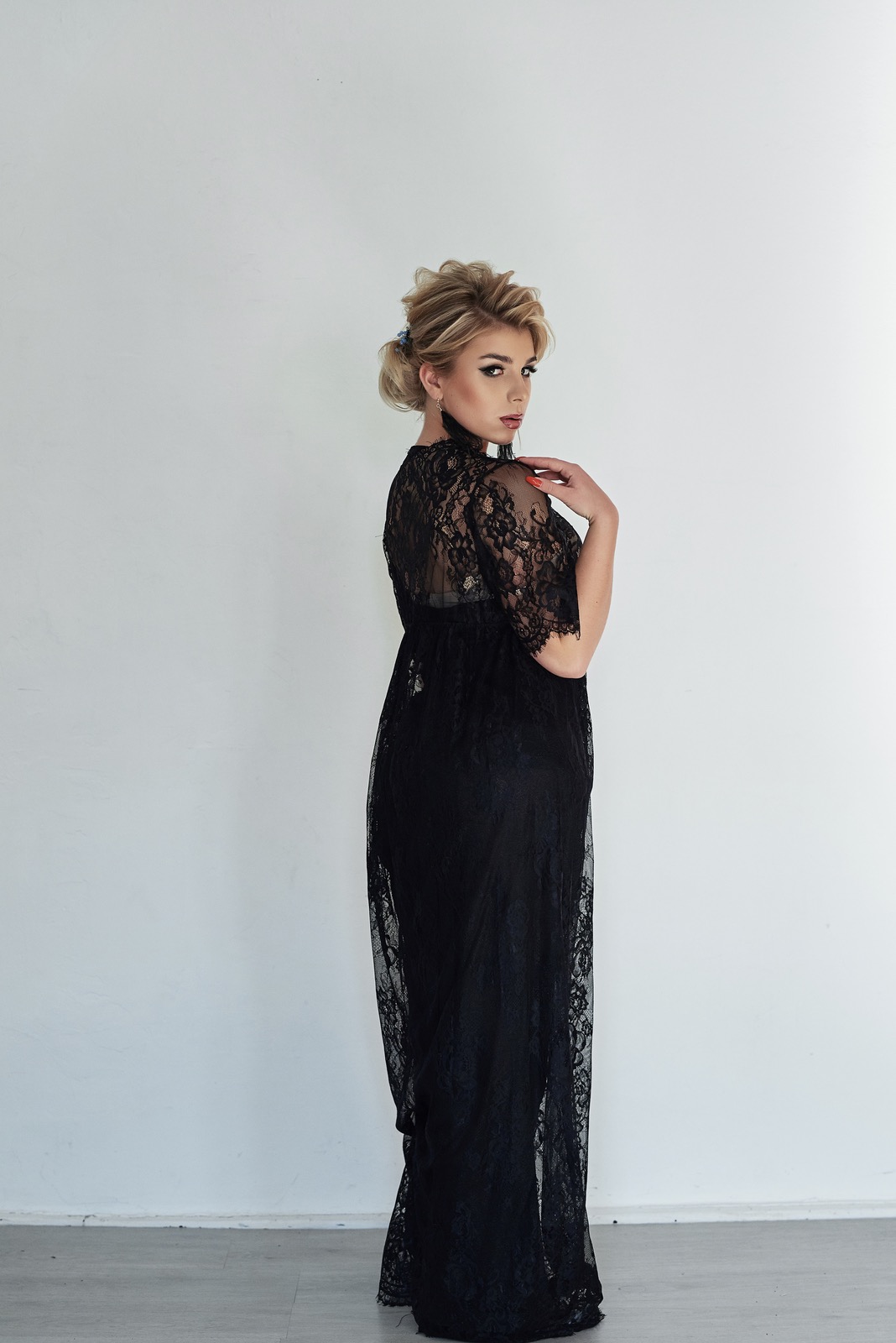 Kobieta w czarnej, koronkowej sukni, odwrócona lekko przez ramię, z upiętymi blond włosami i wyrazistym makijażem, pozuje na tle białej ściany.