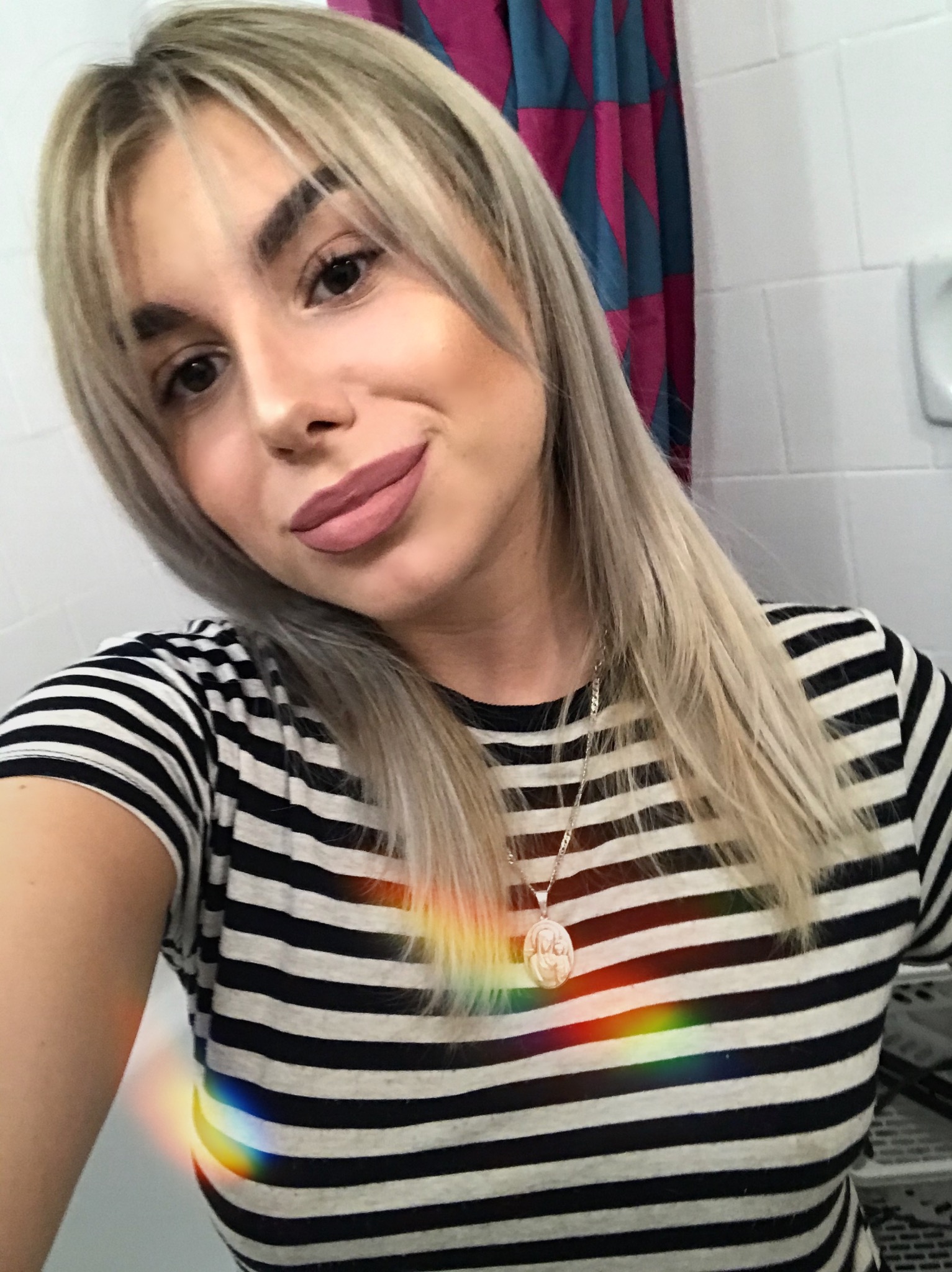 Portret uśmiechniętej kobiety z blond włosami i grzywką, ubrana w koszulkę w czarno-białe paski, z delikatnym makijażem i naszyjnikiem, z tęczowym efektem świetlnym na koszulce.