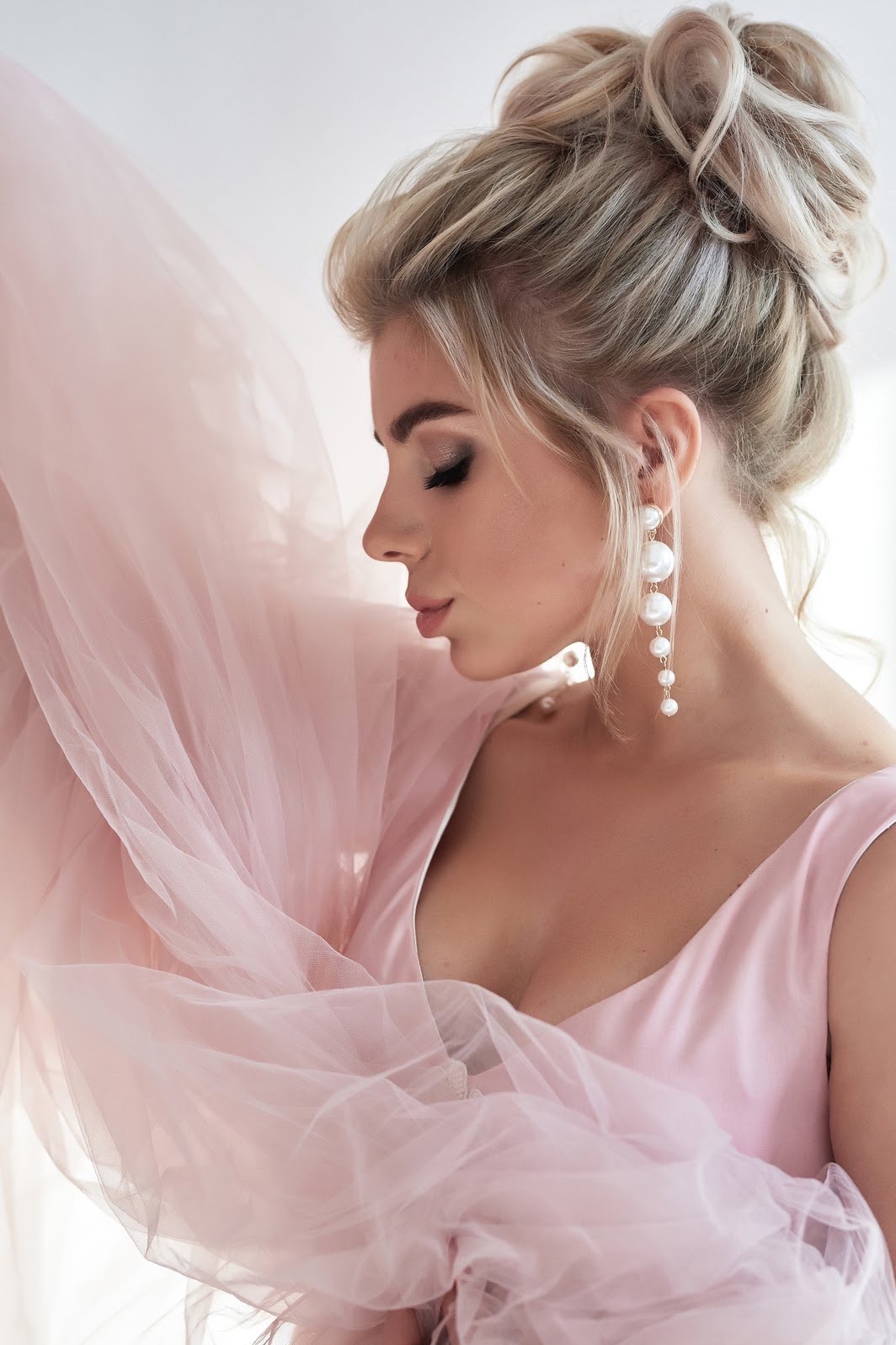 Portret kobiety z zamkniętymi oczami w różowej sukience, z upiętymi blond włosami i długimi perłowymi kolczykami, otulona delikatnym różowym tiulem.