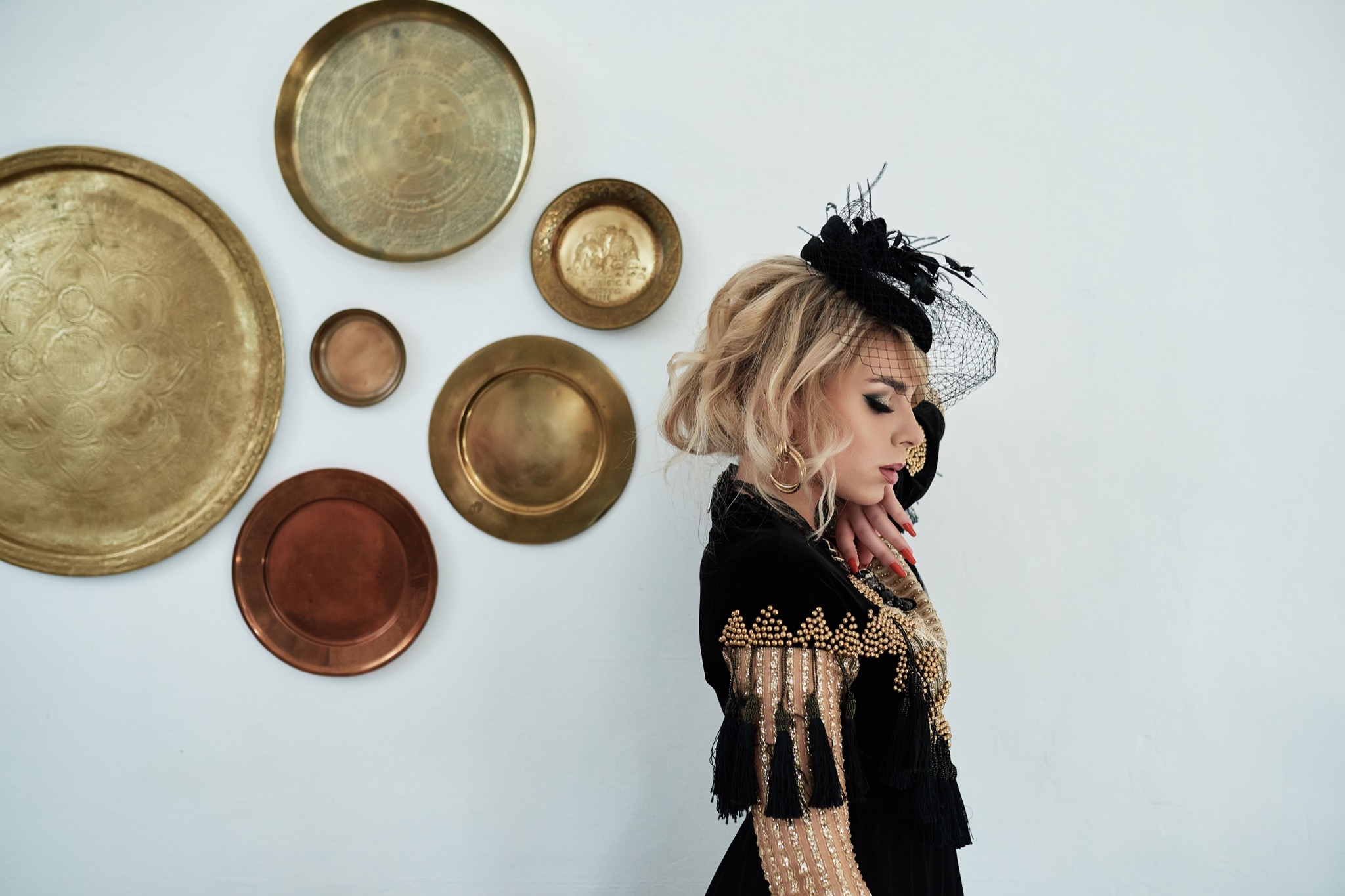 Portret kobiety w profilu, w czarnej sukni z frędzlami i kapeluszu z woalką, pozującej na tle białej ściany z dekoracyjnymi talerzami.