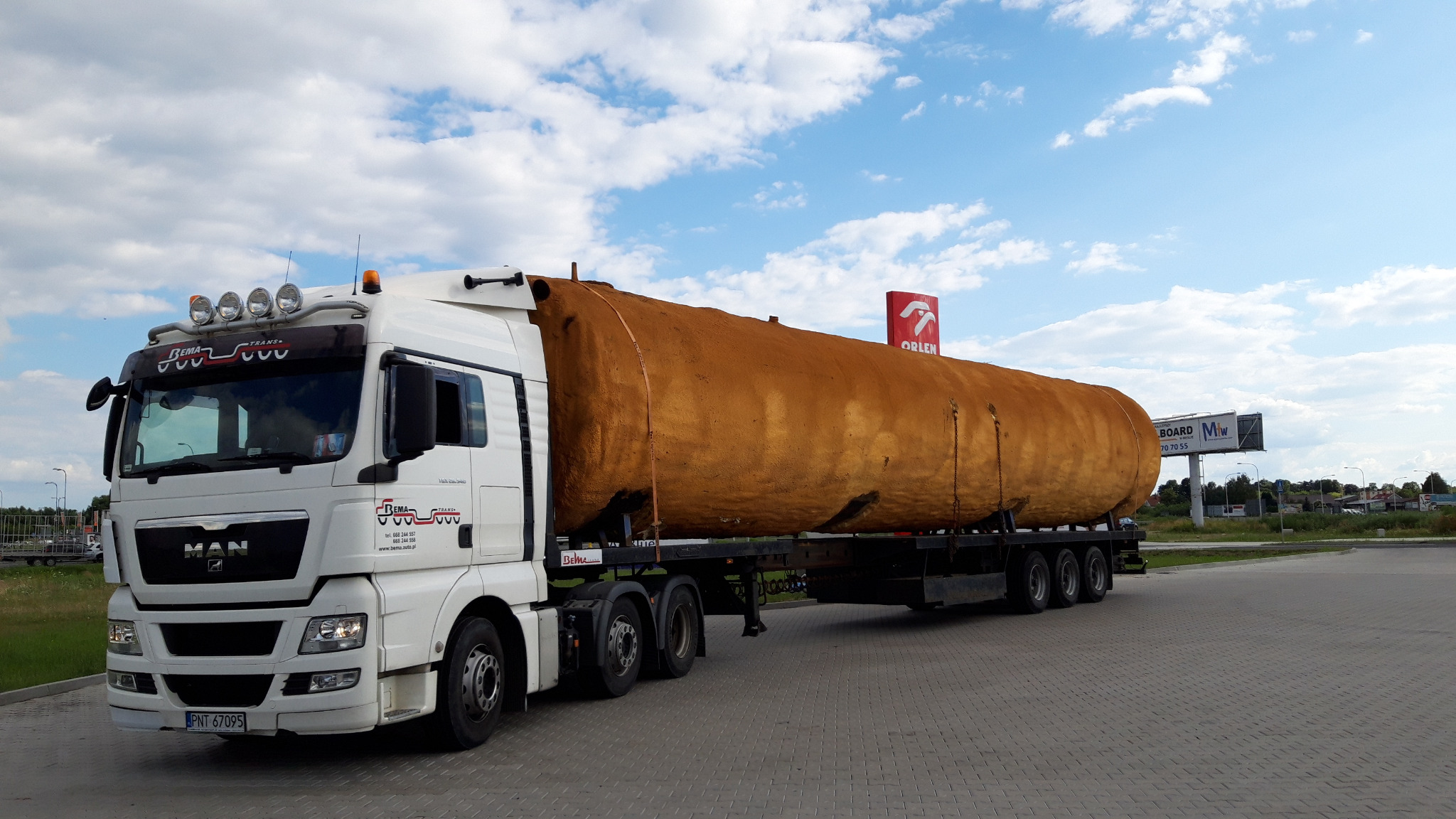 Biała ciężarówka MAN z naczepą przewożąca duży, rdzawy zbiornik cysternę na tle błękitnego nieba z obłokami i znaku stacji Orlen.