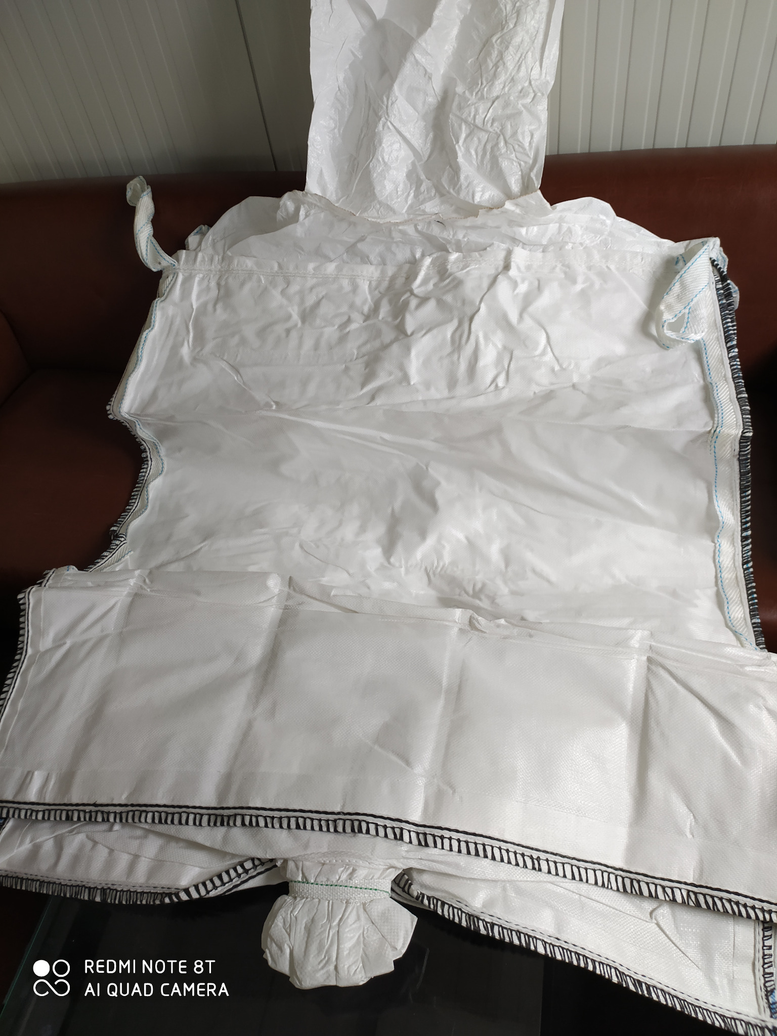 Duży, biały worek Big Bag z polipropylenu, widoczne szwy i uchwyty, leżący na brązowej sofie.