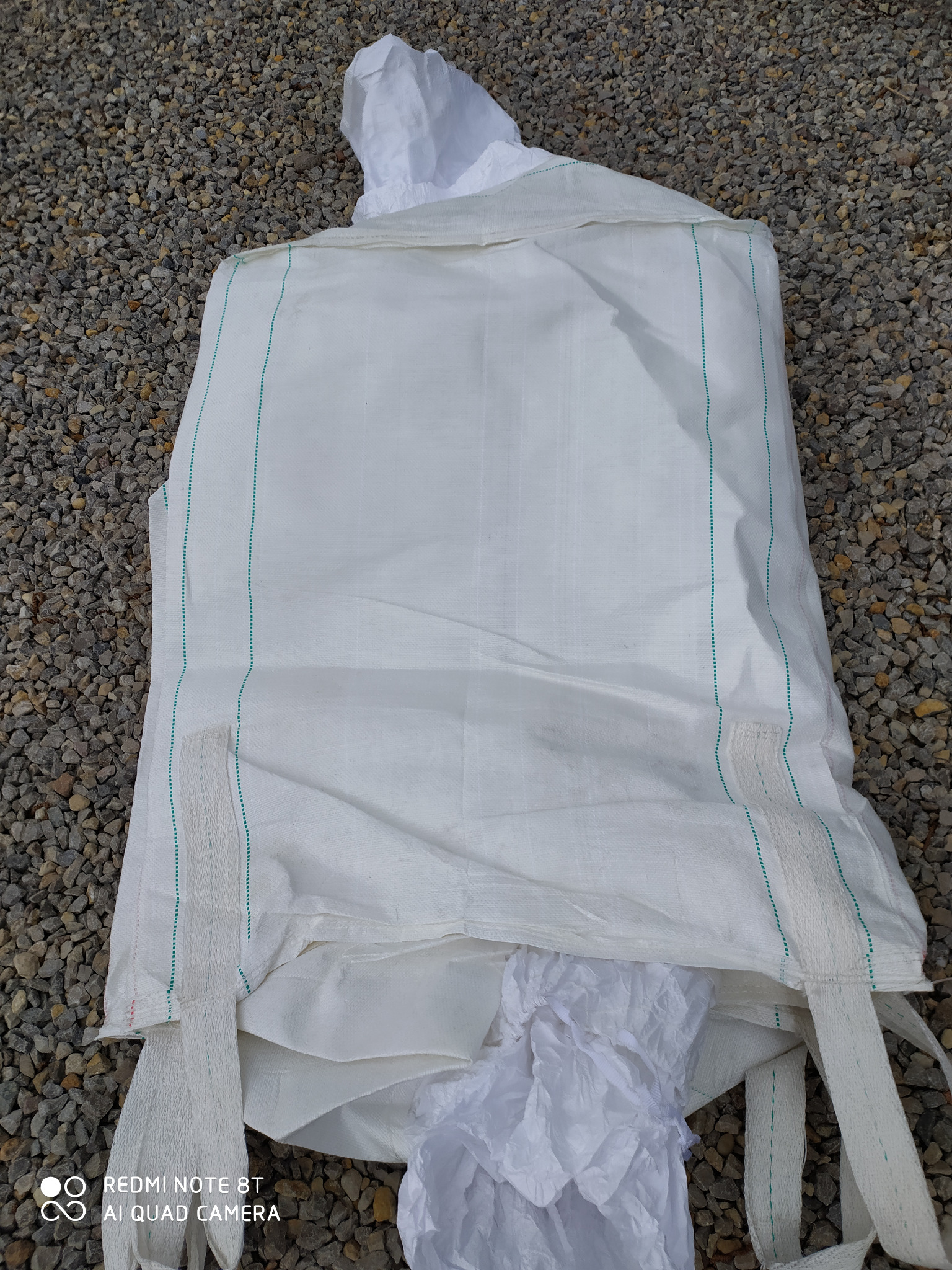 Duży biały worek z tkaniny technicznej z zielonymi przeszyciami, leżący na szarym żwirze, częściowo wypełniony białym materiałem.