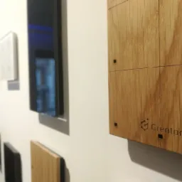 Panel sterowania inteligentnego domu z drewnianą obudową i logo Grenton, zamontowany na ścianie obok innych paneli o różnych kolorach i wykończeniach.