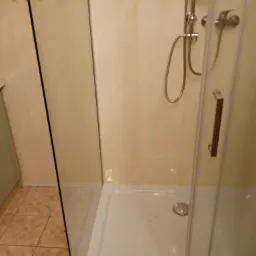 Kabina prysznicowa z brodzikiem i chromowaną armaturą na tle beżowych ścian, widoczne płytki podłogowe.