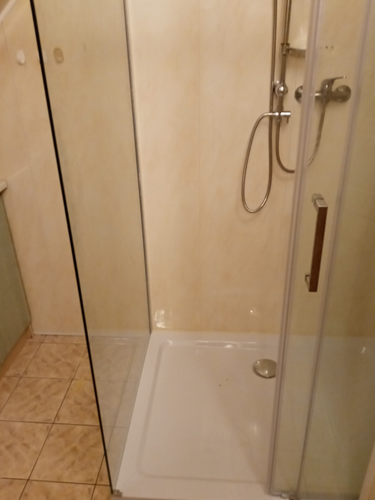 Kabina prysznicowa z brodzikiem i chromowaną armaturą na tle beżowych ścian, widoczne płytki podłogowe.