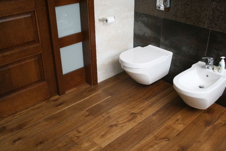 Wykończona łazienka z dębową podłogą, widok na toaletę podwieszaną, bidet i fragment drzwi z szybą.