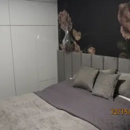 Sypialnia z szarą tapicerowaną ramą ł&oacute;żka, poduszkami i narzutą w odcieniach szarości, na ścianie fototapeta z motywem kwiatowym, obok biała szafa z połyskiem i wisząca lampa w kształcie kropli.