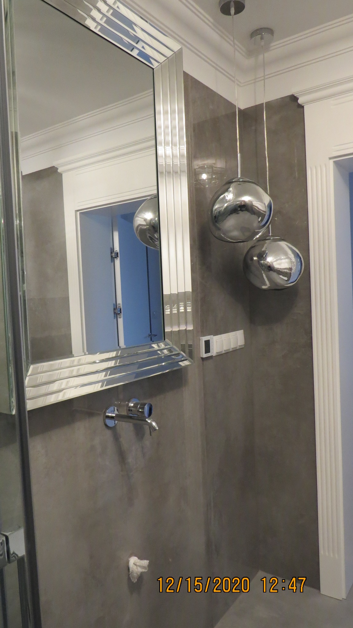 Nowoczesna łazienka z szarą ścianą, lustrem w ozdobnej ramie, chromowaną baterią ścienną i dwoma wiszącymi lampami w kształcie kul.