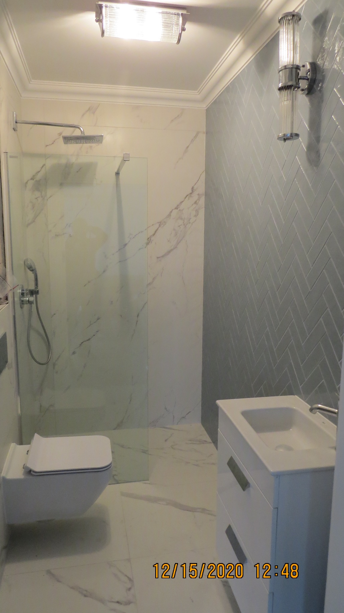 Nowoczesna łazienka z białą armaturą, marmurowymi płytkami i szarą ścianą w jodełkę, oświetlona przez lampę sufitową i kinkiet z kryształkami.