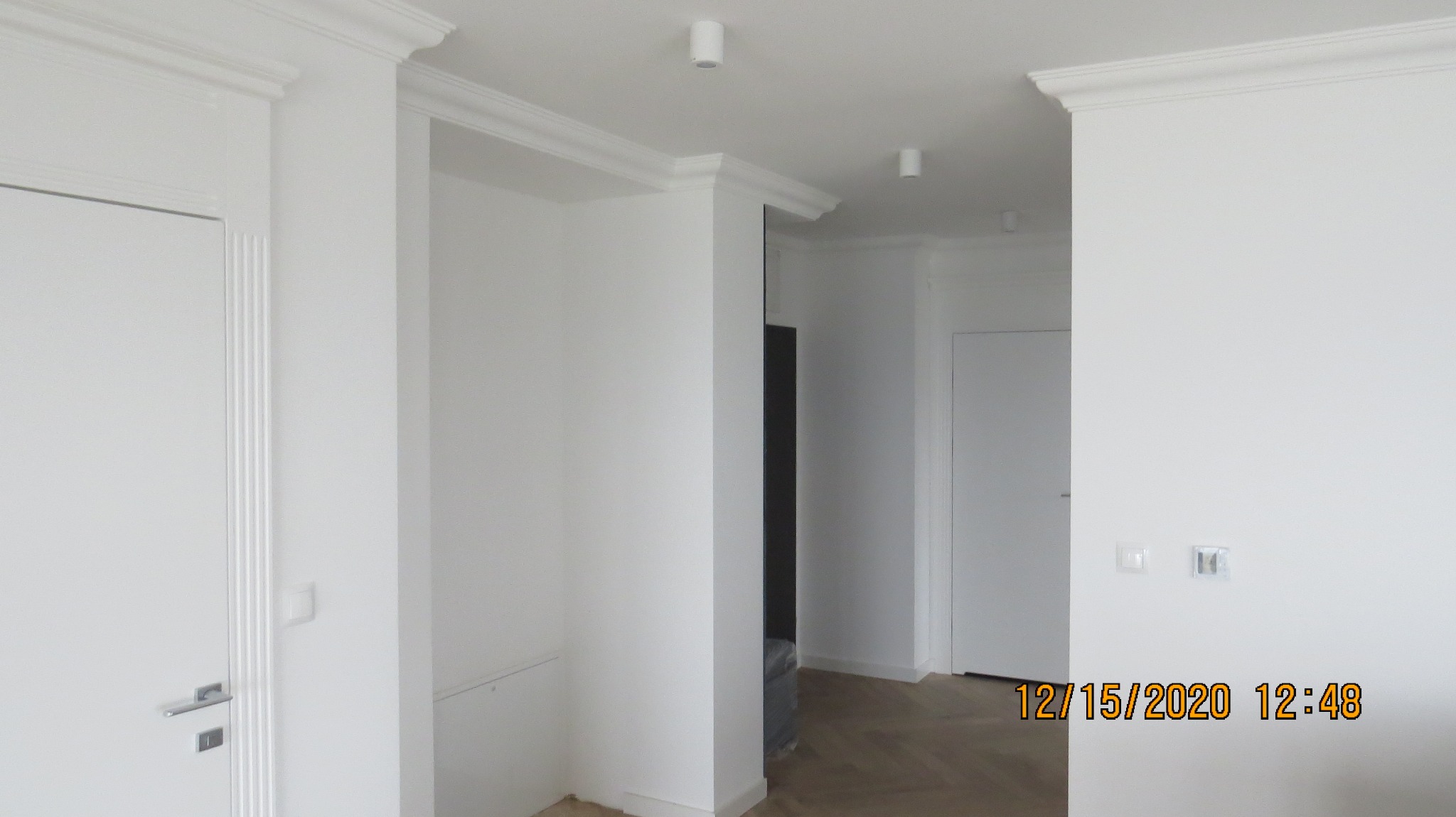 Jasne, nowocześnie wykończone wnętrze z białymi ścianami, sztukaterią sufitową i ozdobnymi listwami wokół drzwi, parkiet w jodełkę na podłodze.