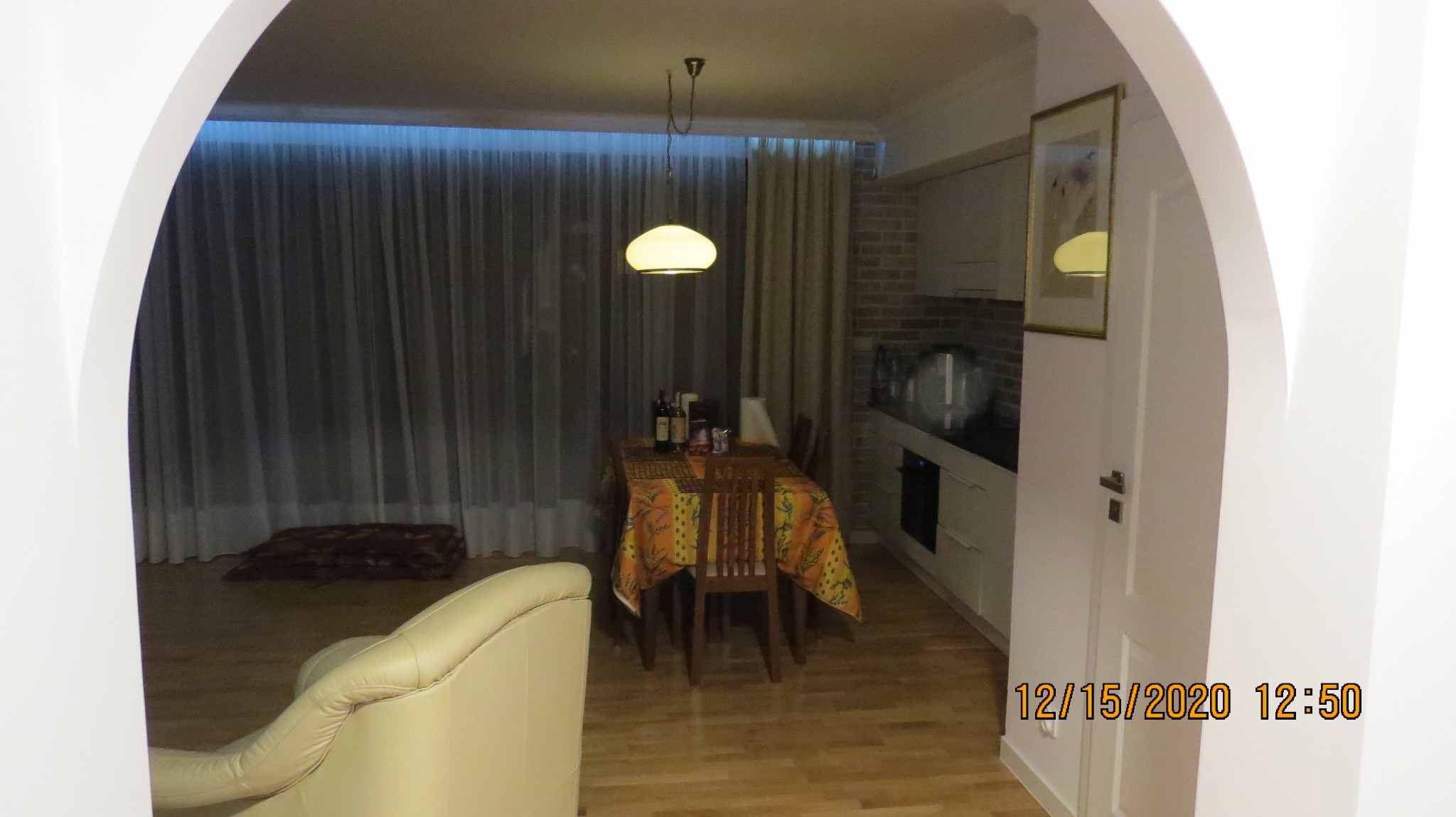 Widok przez łuk na jadalnię z kuchnią w tle, z widoczną lampą wiszącą, zasłonami i fragmentem sofy.