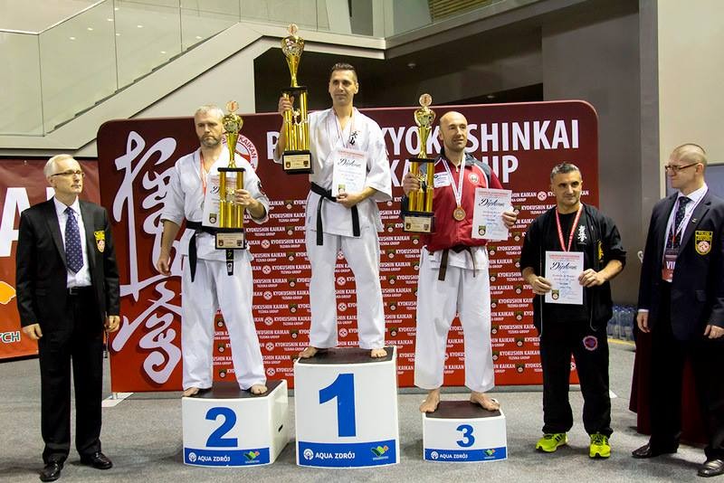 Zawodnicy sportów walki na podium po turnieju Kyokushin Karate, trzymają trofea i dyplomy, widoczny baner z logo Kyokushin Kaikan.