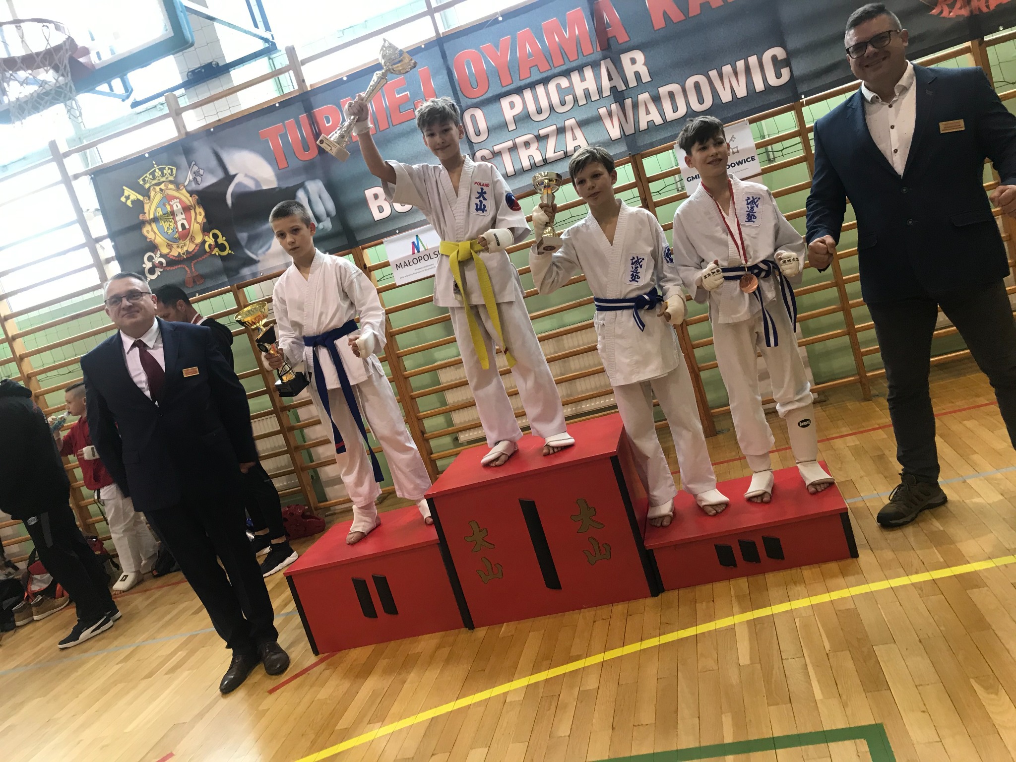 Grupa młodych zawodników w kimonach na podium po turnieju sztuk walki, trzymających trofea, z trenerami w tle.