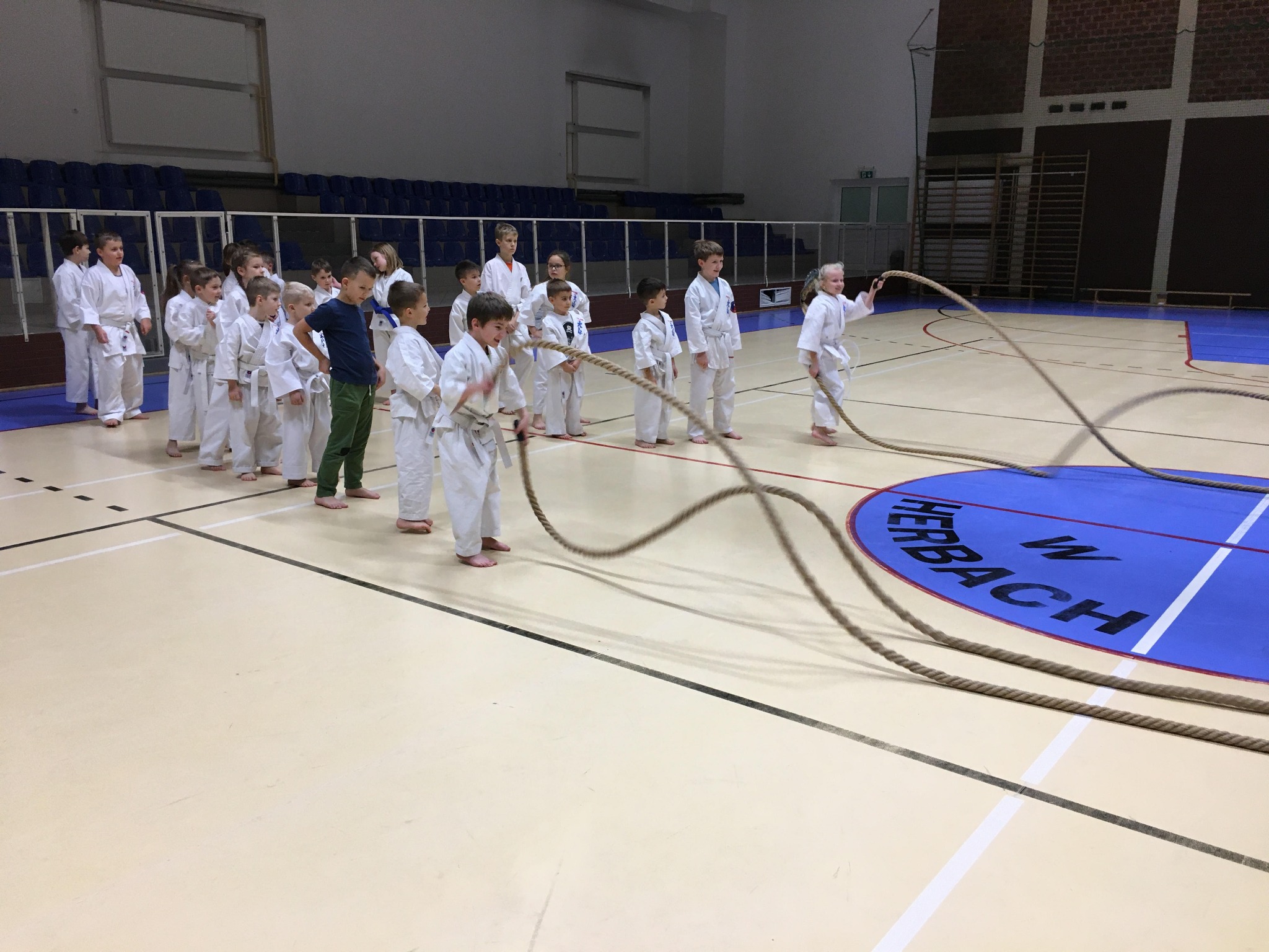 Grupa dzieci w białych kimonach ćwiczy z liną w hali sportowej, na podłodze widoczne logo klubu.