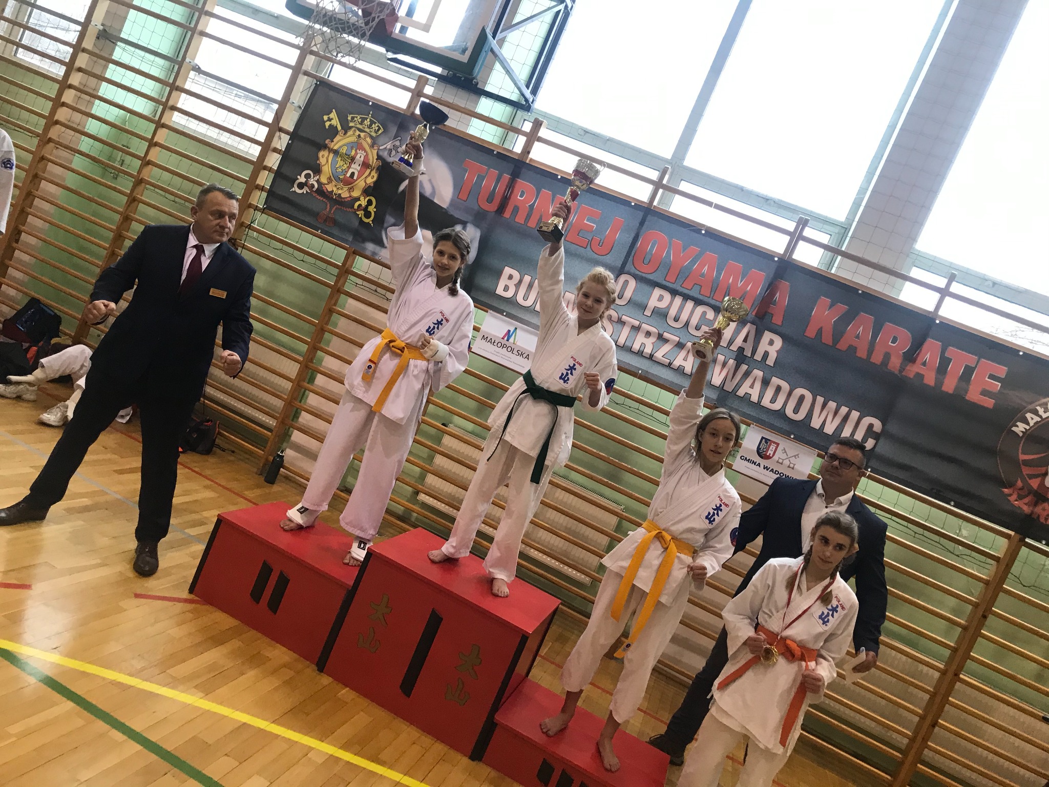 Grupa młodych zawodników karate w białych kimonach z różnymi kolorami pasów na podium, trzymających trofea po turnieju Oyama Karate, na tle baneru reklamowego.