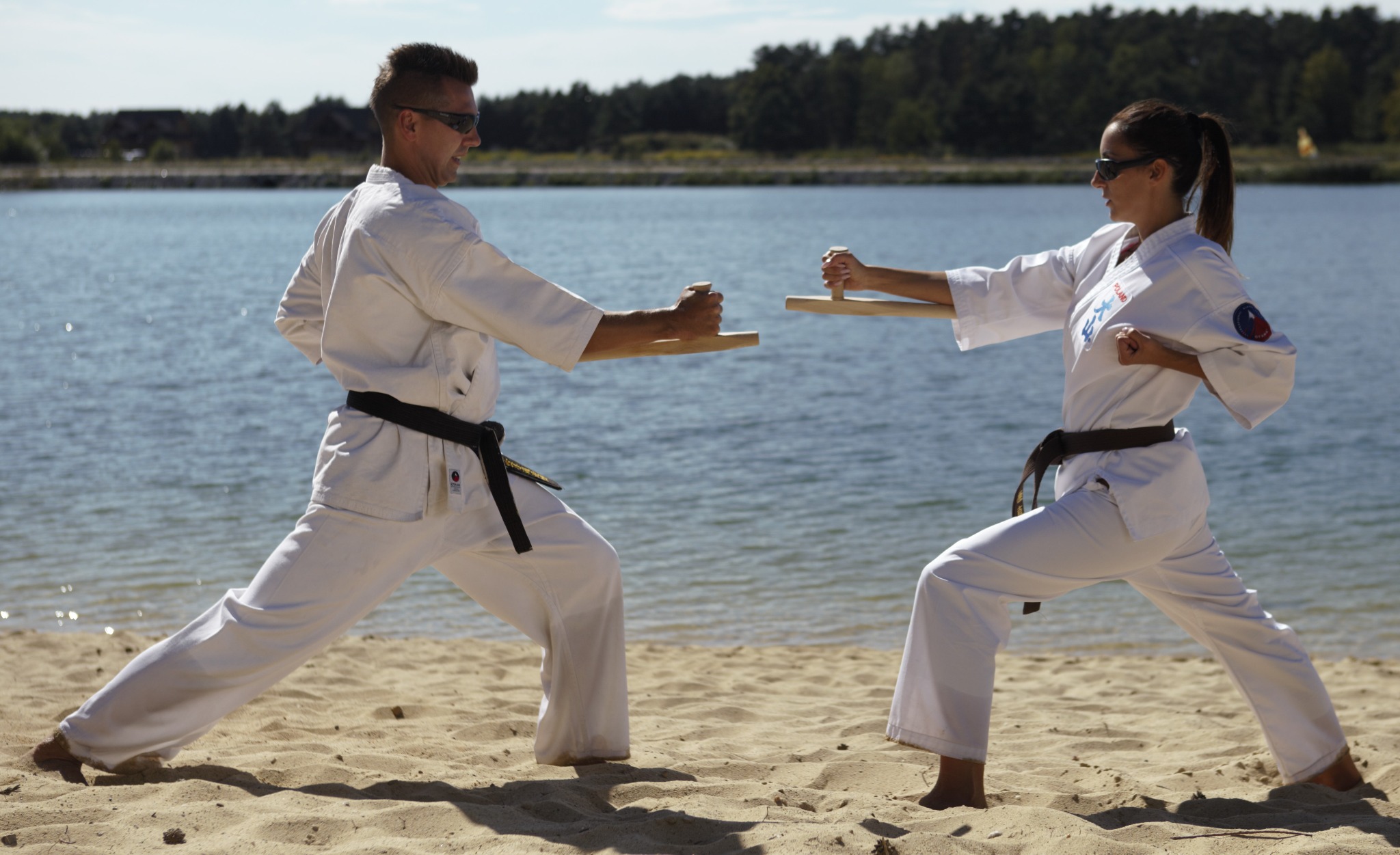 Dwoje ćwiczących karate na plaży, z deskami do łamania, w tle woda i las.