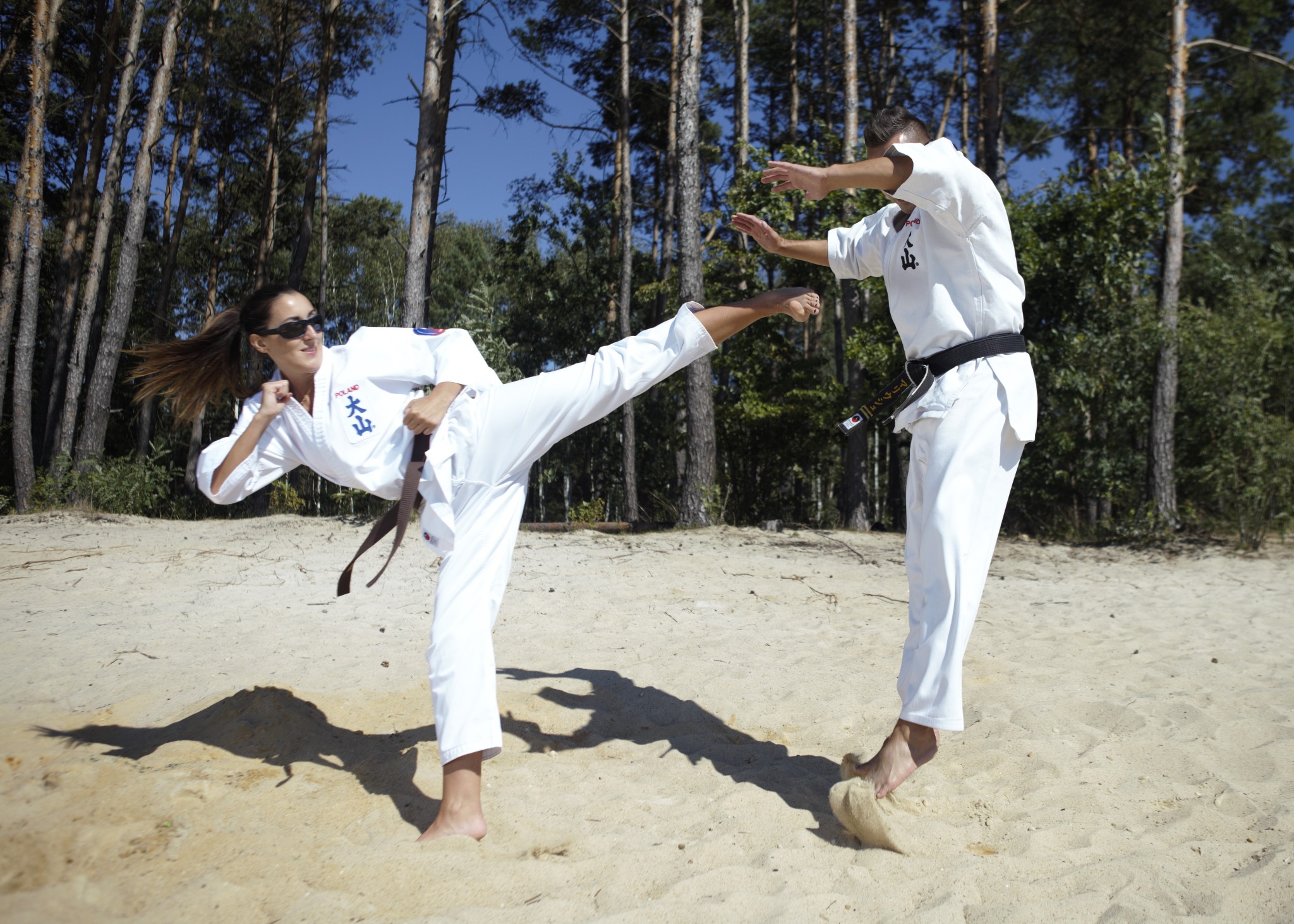 Kobieta w białym karate-gi z brązowym pasem wykonuje kopnięcie boczne w kierunku partnera na piaszczystej plaży, w tle las sosnowy i błękitne niebo.