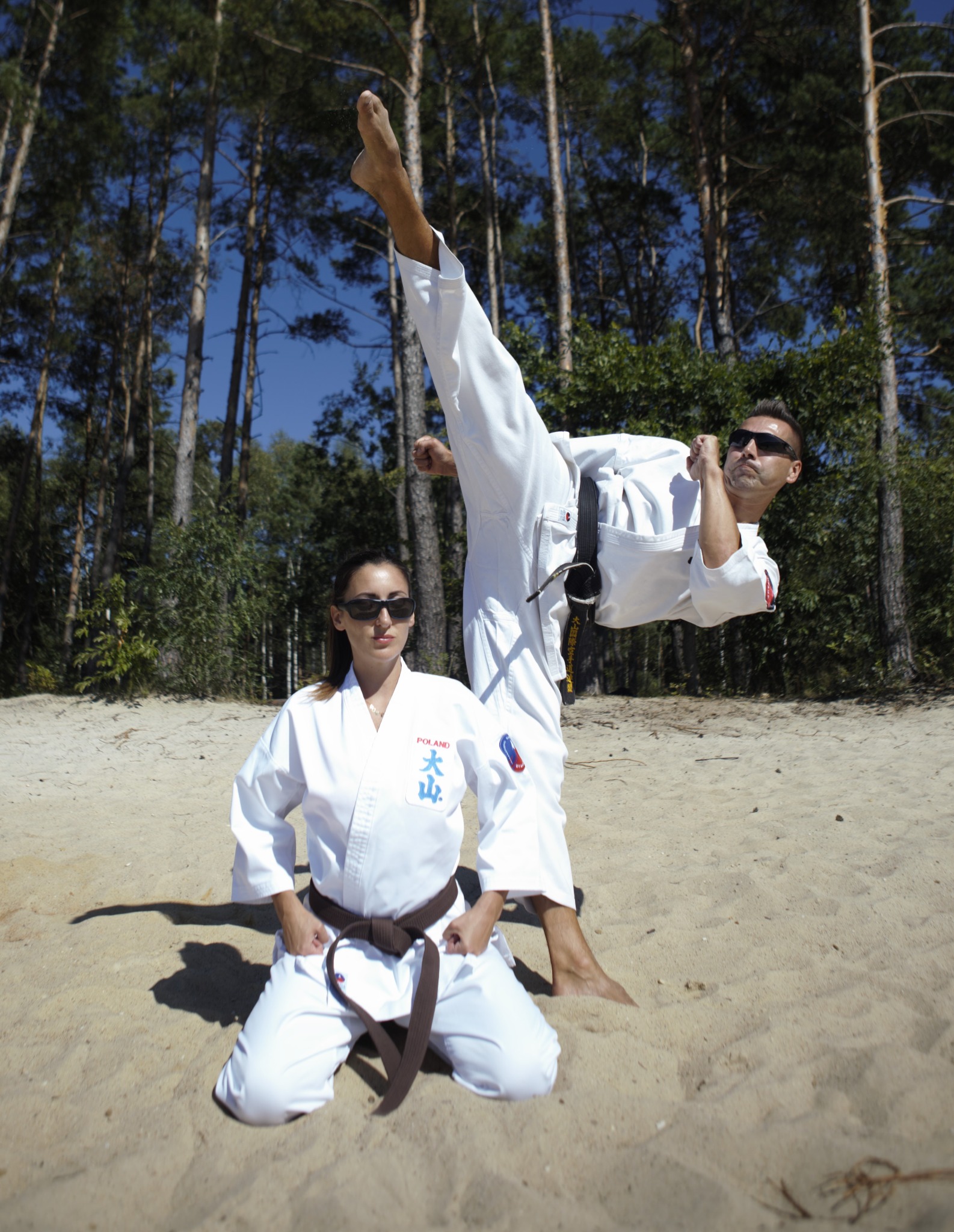 Dwoje ćwiczących karate na piaszczystej plaży w lesie: mężczyzna w okularach wykonuje wysokie kopnięcie, kobieta klęczy z brązowym pasem.