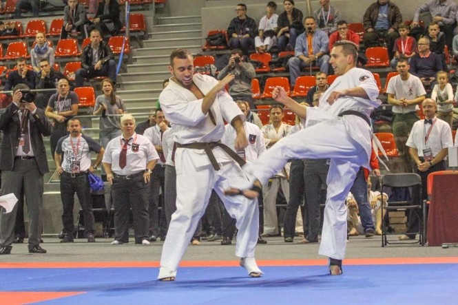 Dwóch mężczyzn w białych strojach karate walczy na tle trybun pełnych widzów podczas zawodów sportów walki.