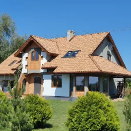 Dom jednorodzinny w trakcie budowy, z widoczną konstrukcją dachu pokrytą dachówką ceramiczną, oknami dachowymi i kominem, elewacja częściowo ocieplona styropianem, otoczony zielenią.