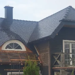 Częściowo pokryty łuską dach domu z drewnianą elewacją, widoczne rusztowania i fragmenty starej konstrukcji dachu, antena satelitarna na bocznej ścianie.