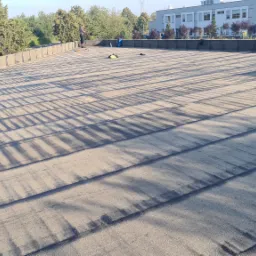 Rozległy, płaski dach pokryty papą bitumiczną z widocznymi cieniami, pracownik z narzędziami w oddali, budynek w tle.