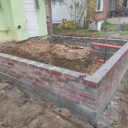 Murowana podmurówka z czerwonej cegły, częściowo wypełniona ziemią, z widocznymi pomarańczowymi rurami i rękawicami roboczymi na krawędzi.