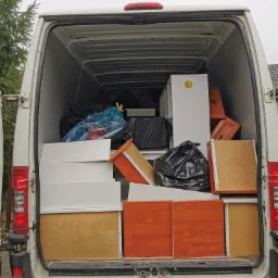 Wnętrze białego busa załadowane meblami i workami z rzeczami podczas transportu, widoczna naklejka z uśmiechniętą buźką na białej szafce.