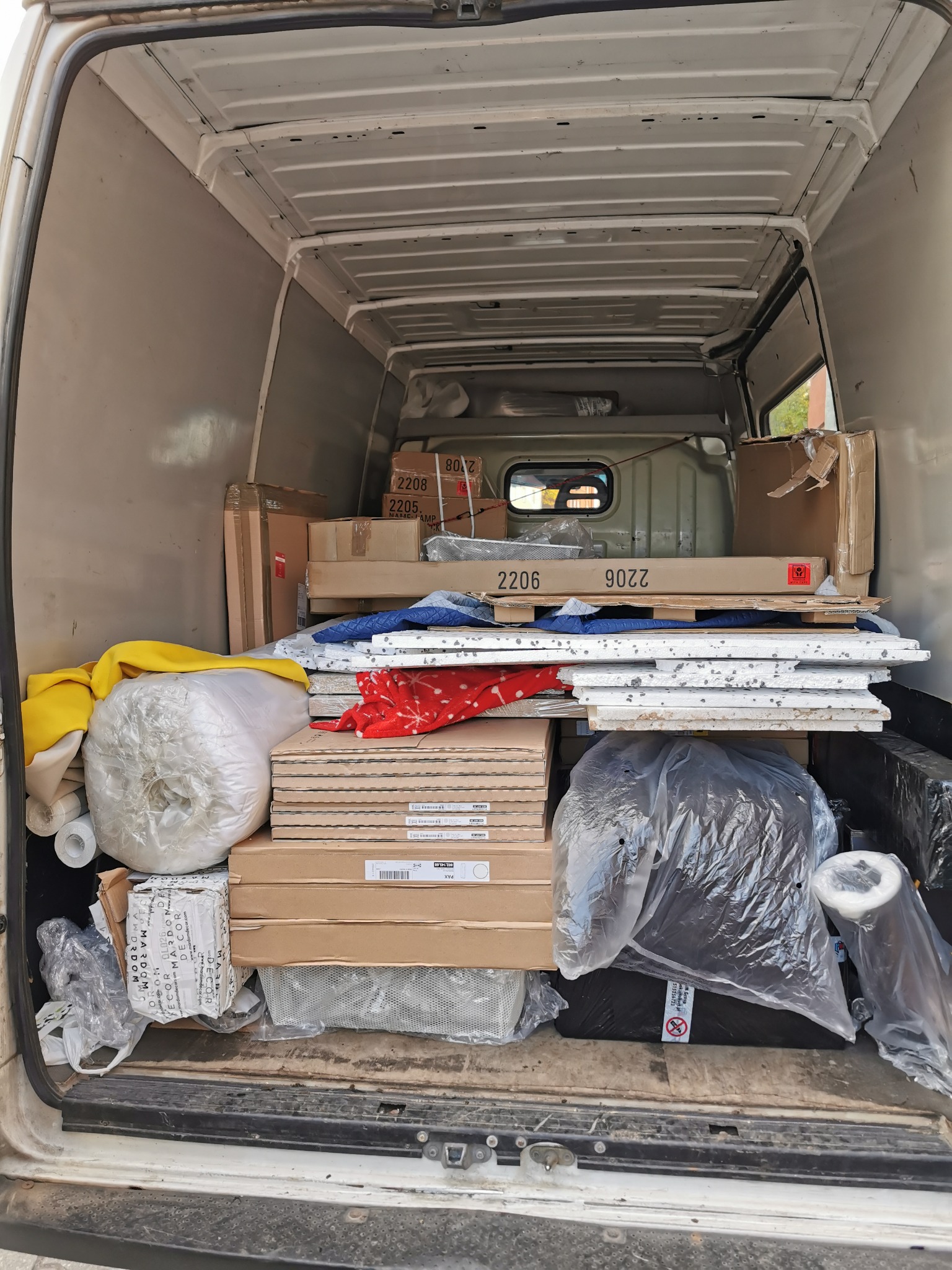 Wnętrze białego busa wypełnione kartonami, rolkami materiałów i elementami wyposażenia wnętrz, widoczne oznaczenia '2206' i '2205' na kartonach.