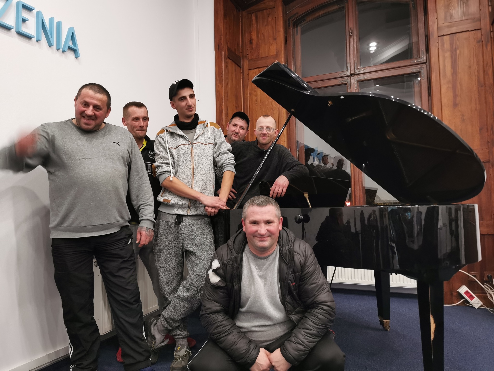 Grupa sześciu mężczyzn pozuje obok czarnego fortepianu w pomieszczeniu z drewnianą boazerią i napisem na ścianie.