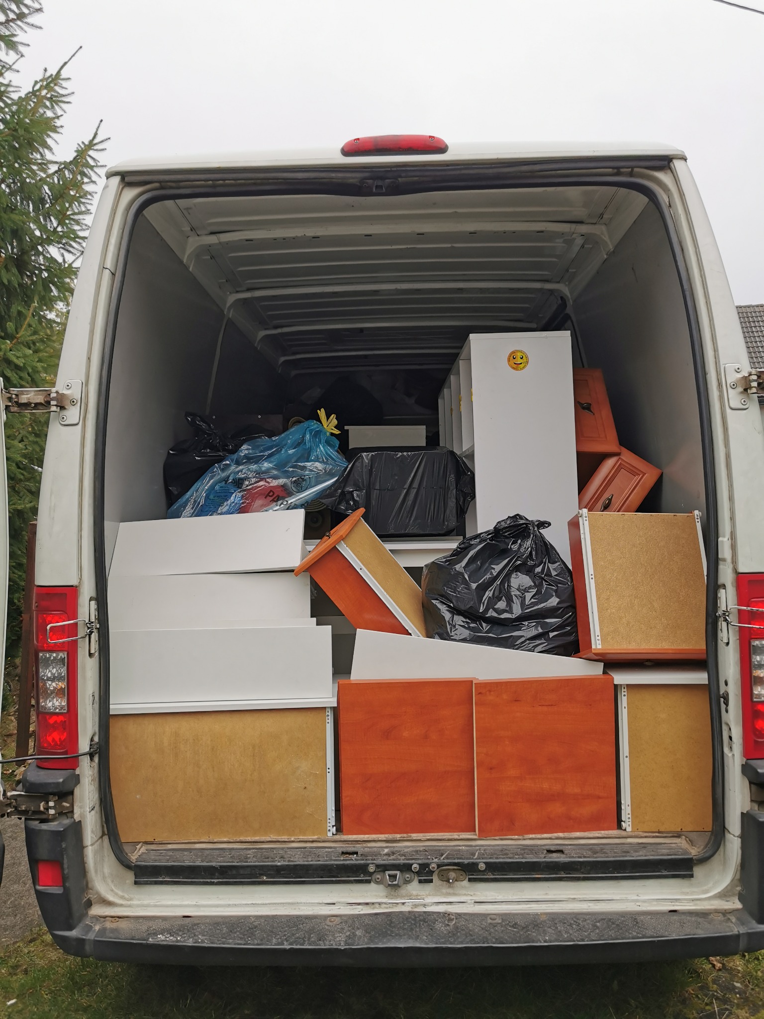 Wnętrze białego busa załadowane meblami i workami z rzeczami podczas transportu, widoczna naklejka z uśmiechniętą buźką na białej szafce.