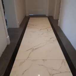 Korytarz z nowo położonymi płytkami podłogowymi w odcieniach bieli i szarości, z ciemnoszarym obramowaniem, prowadzący do drzwi w głębi.