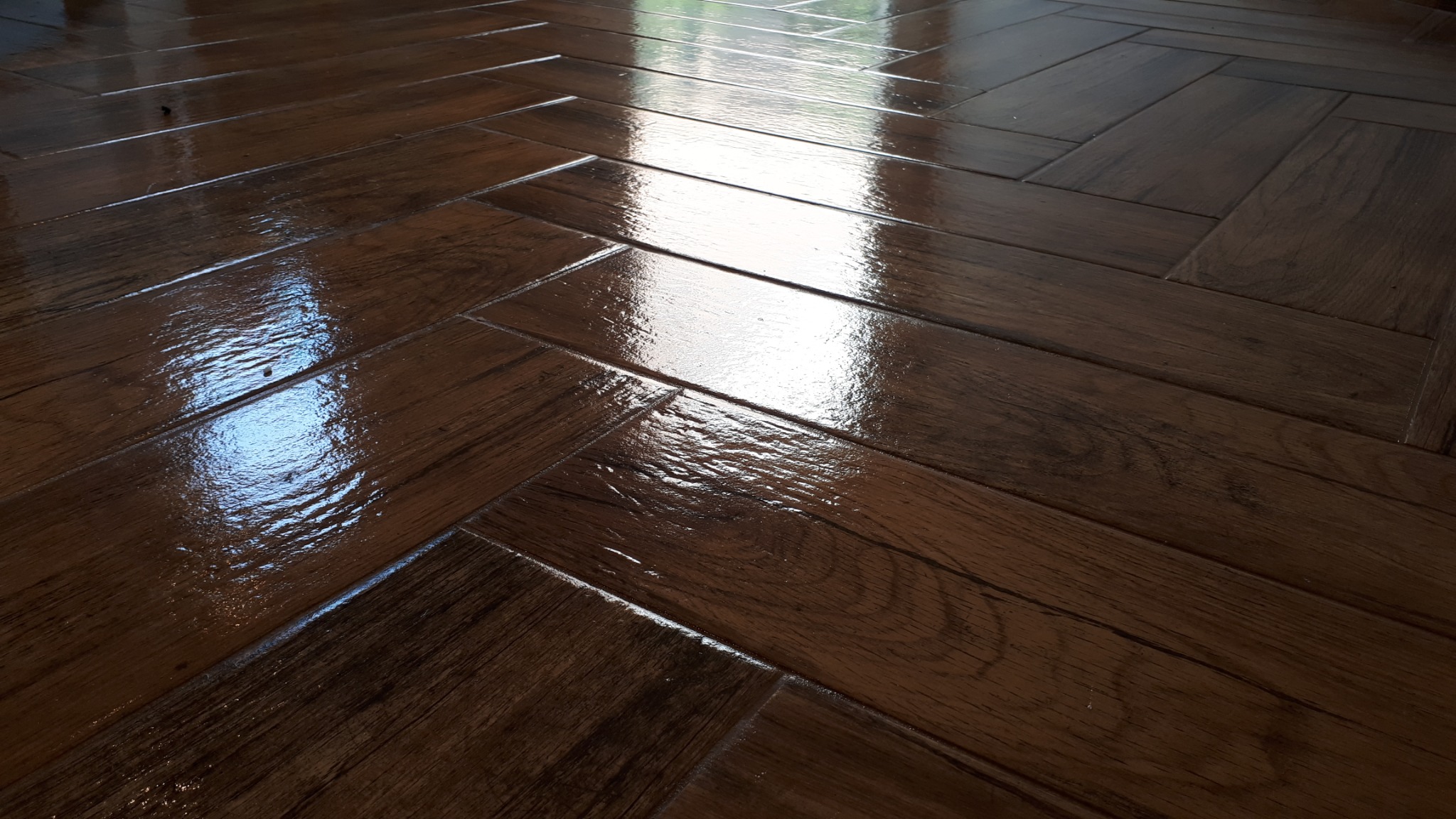 Błyszcząca, ciemnobrązowa podłoga ułożona w jodełkę, odbijająca światło okna, imitująca drewno.