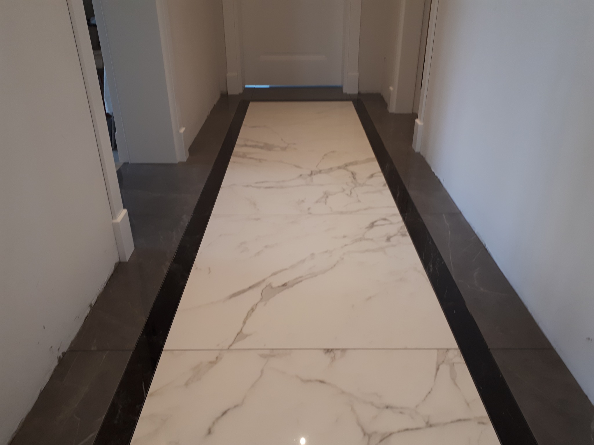 Korytarz z nowo położonymi płytkami podłogowymi w odcieniach bieli i szarości, z ciemnoszarym obramowaniem, prowadzący do drzwi w głębi.