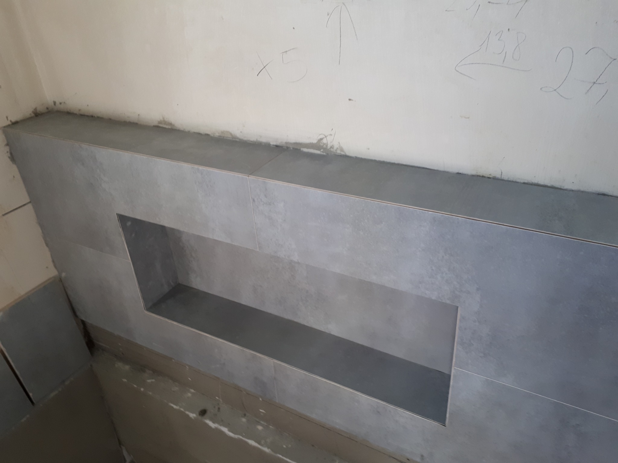 Półka wnękowa w ścianie wyłożona szarymi płytkami ceramicznymi, widoczne niedoskonałości i zaprawione fugi na ścianie, surowy stan wykończenia, ślady ołówka na ścianie powyżej.