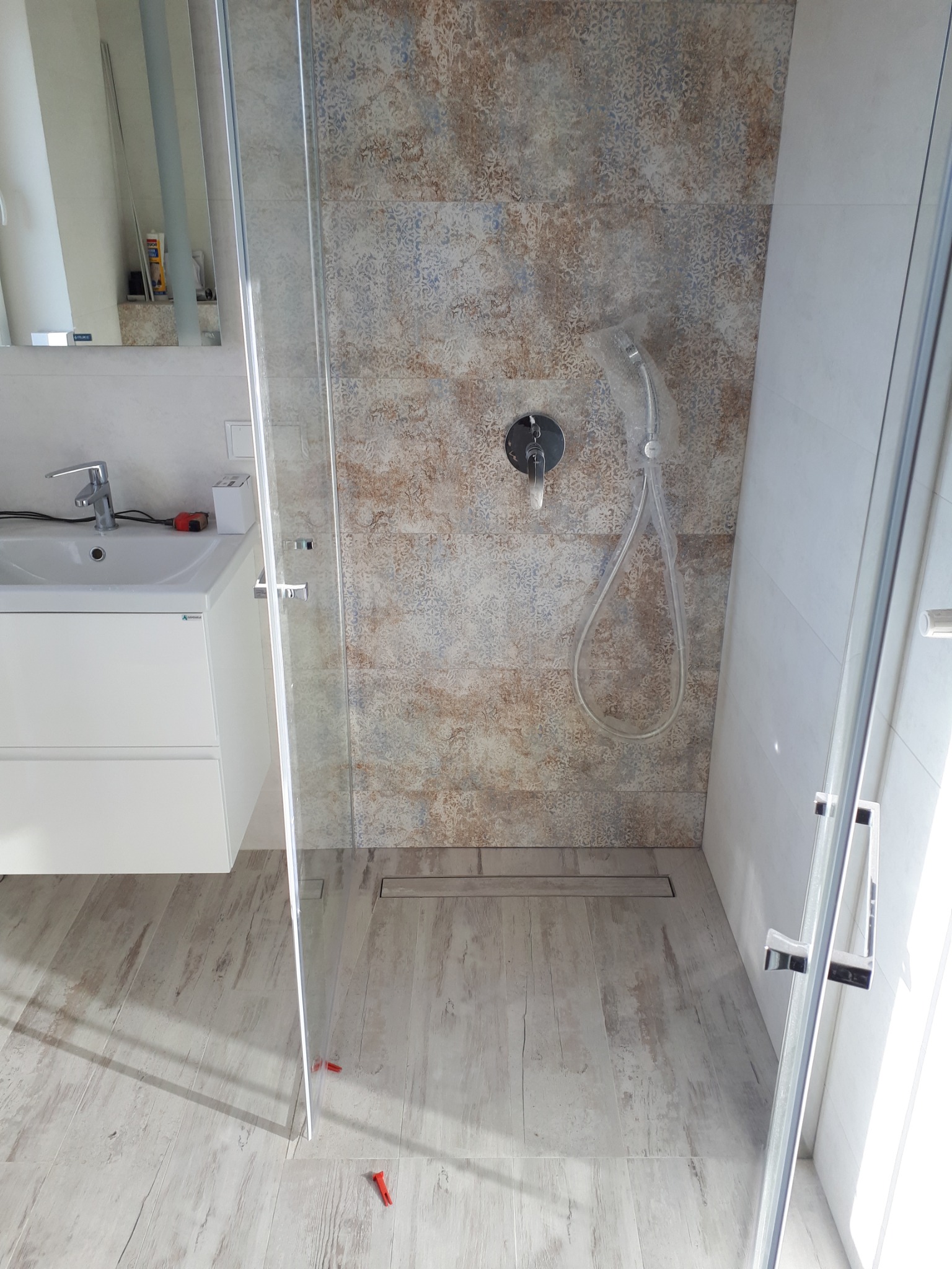 Nowoczesna łazienka z kabiną prysznicową typu walk-in, wykończona płytkami w odcieniach beżu i błękitu, z odpływem liniowym w podłodze imitującej drewno. Bateria prysznicowa z deszczownicą w folii...